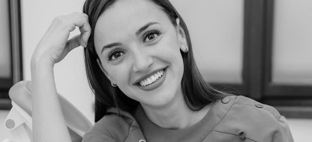 Fotografie alb-negru: Cristina Păcurar în cabinet, sprijinită de tetiera scaunului, cu mâna ridicată în păr, privind spre aparat și zâmbind. În dreapta, ferestrele înalte ale încăperii.